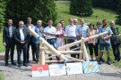 Zahlreiche Personen stehen hinter einer Barriere aus Holz. Davor sind Tafeln mit Wappen und Logos aus Vorarlberg und Bayern platziert.  