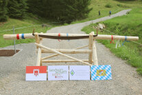 Eine Barriere aus Holz versperrt noch den Alpweg. Vier Tafeln, jeweils mit Wappen oder Logo aus Vorarlberg und Bayern, stehen für die bilaterale Zusammenarbeit.
